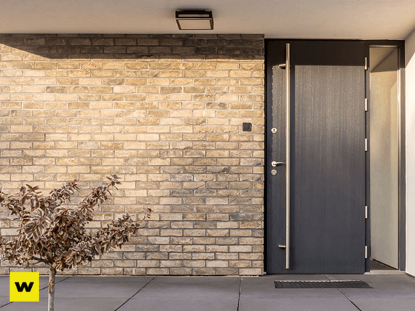 Moderne Haustür aus dunklem Material neben heller Klinkerfassade mit kleinem Baum davor.